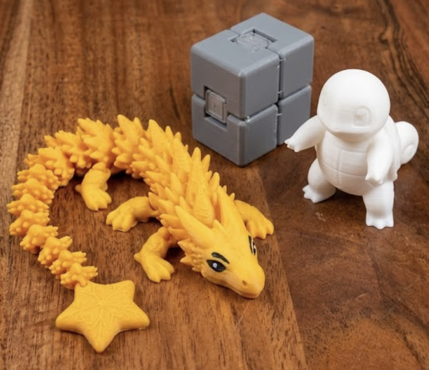 3D-gedruckte Figuren: gelber Gelenkdrache mit Stern, grauer Infinity Cube und weiße Schiggy-Figur auf Holztisch